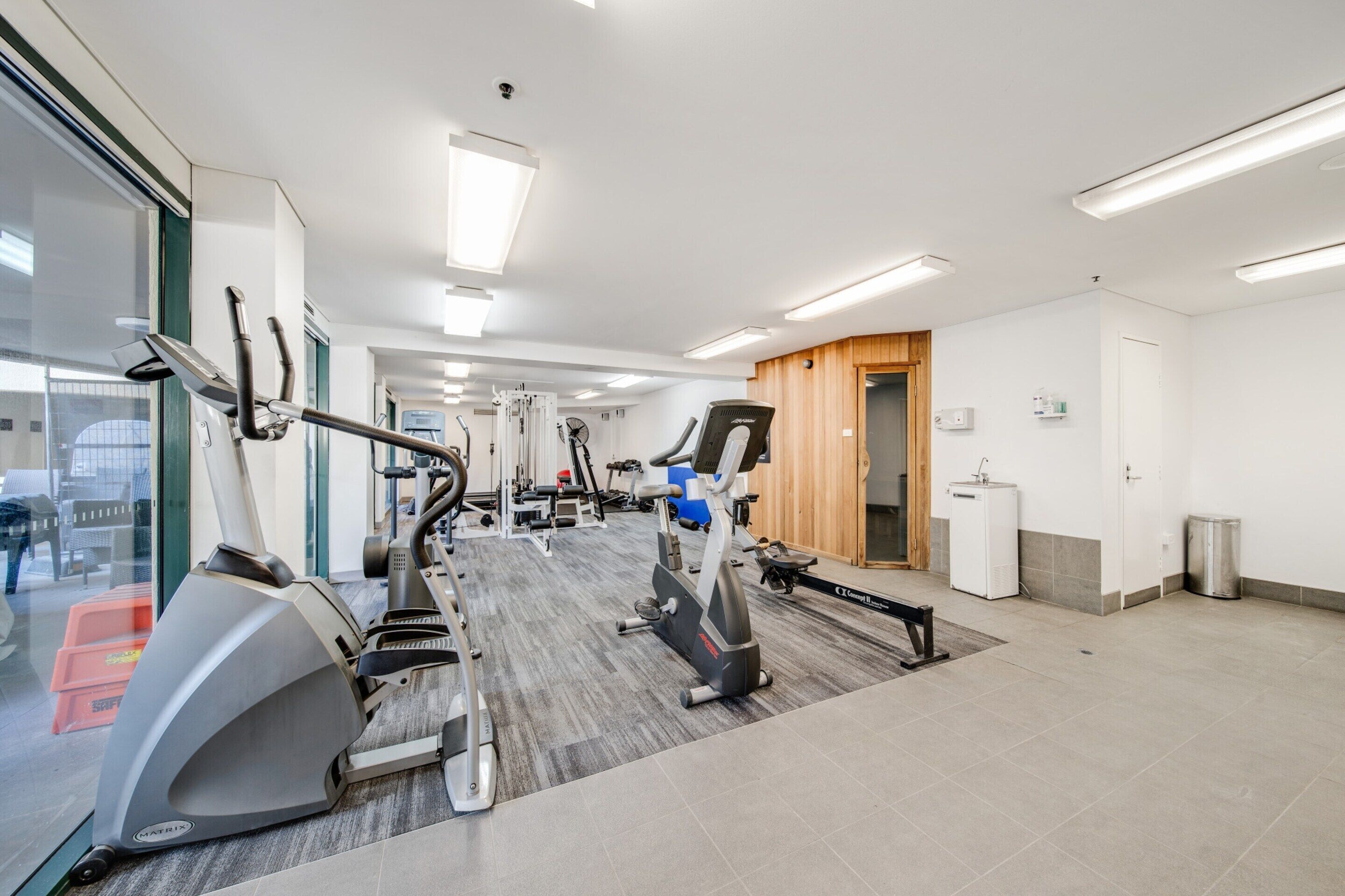 A modern gym with various exercise equipment, a large mirror, and a view of the outside through glass doors.