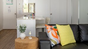 A modern living room with a black leather sofa, a round wicker side table, and a small white vase with a plant. - Seafront Getaway Studio, Great Location With Pool (Batehaven)