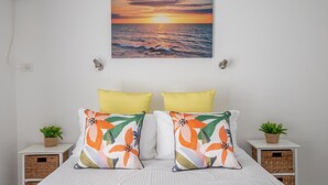 A bedroom with a bed, two pillows, two nightstands with plants, and a sunset painting on the wall. - Seafront Getaway Studio, Great Location With Pool (Batehaven)
