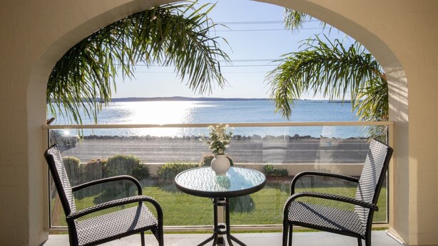 A balcony with two wicker chairs and a glass table, overlooking a body of water and a distant shoreline. - Seafront Getaway Studio, Great Location With Pool (Batehaven)