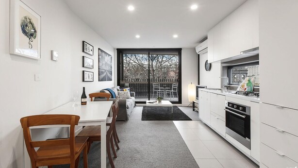 A modern kitchen with white cabinets, a dining area with a wooden table and chairs, and a large window providing a view of the outdoors. - 2 bed Apartment in Trendy Collingwood (Melbourne)