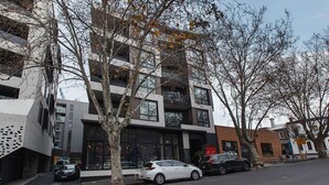 A modern building with a glass facade, parked cars, and leafless trees. - 2 bed Apartment in Trendy Collingwood (Melbourne)