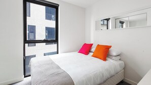 A modern bedroom with a large window, a bed with white bedding and two pillows, one pink and one orange. - 2 bed Apartment in Trendy Collingwood (Melbourne)