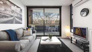 A modern living room with a sofa, coffee table, TV, and a balcony with outdoor furniture. - 2 bed Apartment in Trendy Collingwood (Melbourne)