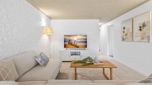 Un salon moderne avec un mur en briques blanches, un canapé, une table basse en bois et un tableau représentant un coucher de soleil sur l’océan.