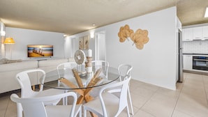 Un salon moderne avec une table à manger à plateau de verre, entourée de chaises blanches, d’un canapé et d’un tableau accroché au mur.