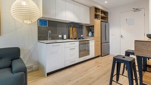 A modern kitchen with white cabinets, a built-in oven, a refrigerator, and a wooden shelf with jars. - City View Haven in South Melbourne (Melbourne)