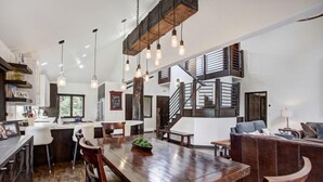 Une cuisine spacieuse avec une table à manger en bois, des tabourets de bar et des luminaires suspendus. Un escalier mène à un niveau supérieur doté d’un balcon.