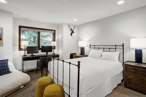 Um quarto com uma cama, duas mesas de cabeceira, uma escrivaninha com cadeira, uma janela e uma decoração de cabeça de veado na parede.