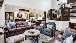 Un salon spacieux avec un canapé en cuir marron, un fauteuil gris et une table basse. Un escalier avec une rampe en bois mène à l’étage supérieur.