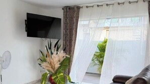 Una sala de estar moderna con un sofá de cuero marrón, una mesa de centro de vidrio, un televisor de pantalla plana montado y un gran ventanal con cortinas traslúcidas.