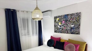 Un dormitorio con una cama, una lámpara colgante, una ventana con cortinas de gasa y un cuadro floral en la pared.