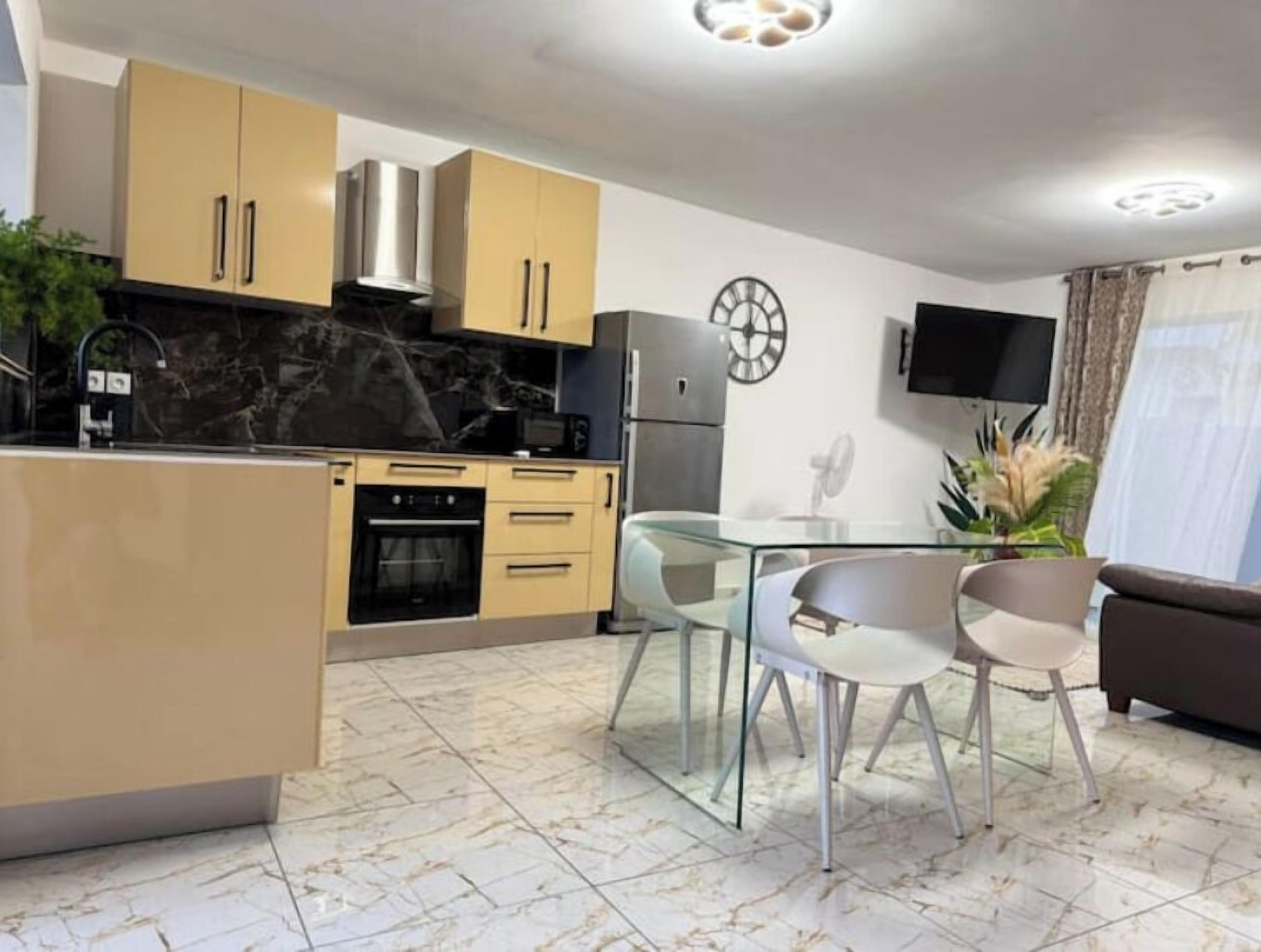 A modern kitchen with beige cabinets, a black countertop, and a marble backsplash. There is a dining area with a glass table and two chairs. A TV is mounted on the wall, and a clock is visible.