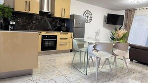 Una cocina moderna con armarios color beige, encimera negra y salpicadero de mármol. Hay una zona de comedor con una mesa de vidrio y dos sillas. Un televisor está montado en la pared, y se observa un reloj.