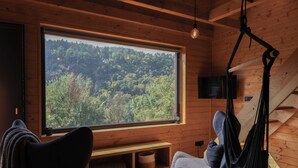 A cozy cabin interior with a large window offering a forest view, a hanging chair, and a bench. - Chata Cedrus (Hradištko)