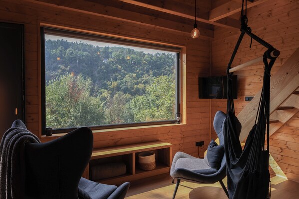 A cozy cabin interior with a large window offering a forest view, a hanging chair, and a bench.