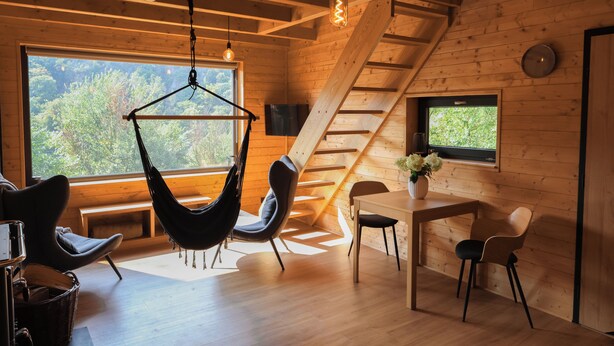 A cozy cabin interior with a wooden interior, a hammock chair, a dining table with chairs, and a large window offering a view of trees. - Chata Cedrus (Hradištko)