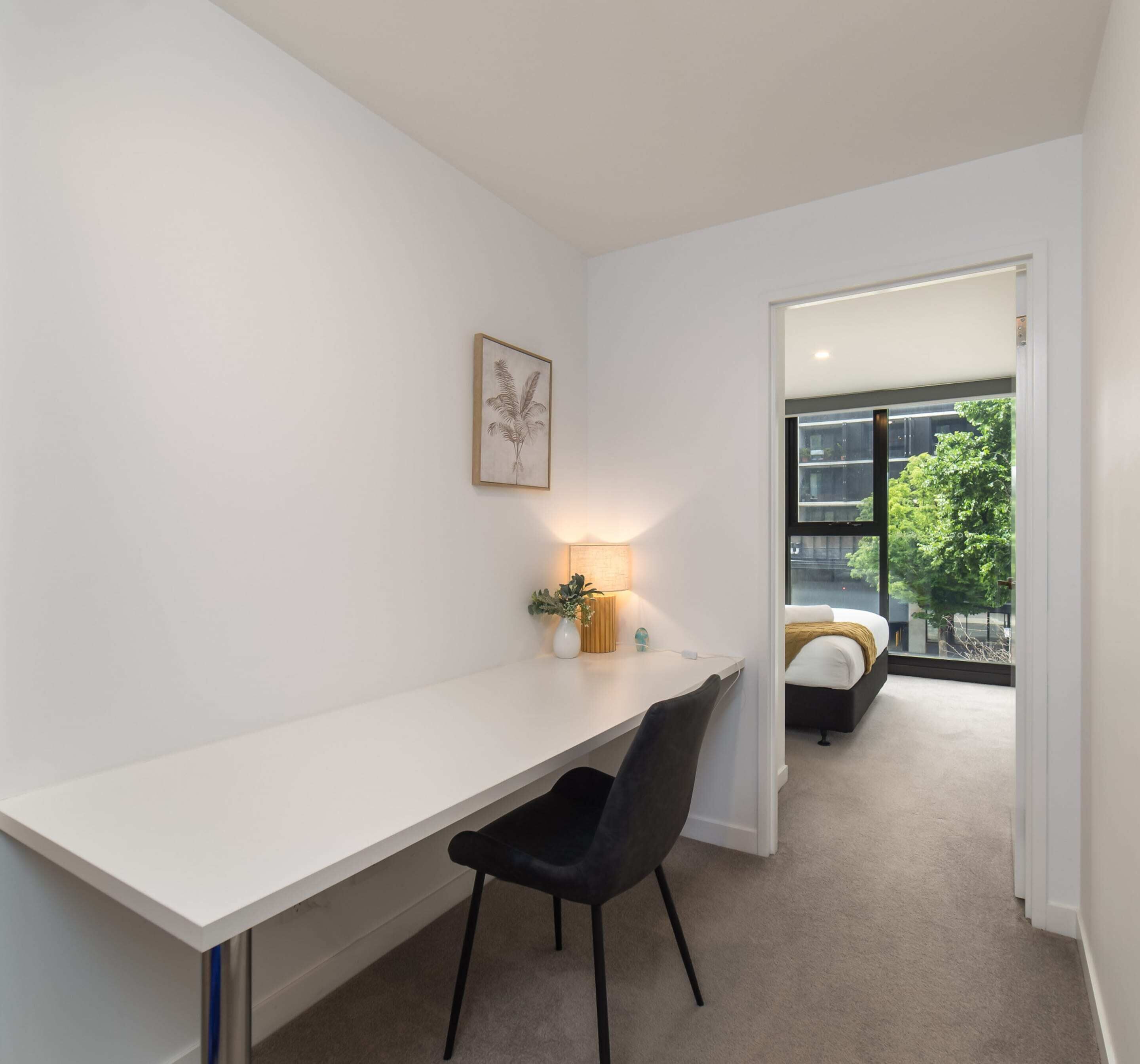 A modern room with a white desk, a black chair, a framed artwork, a bedside table with a lamp, and a view of a balcony with a bed.