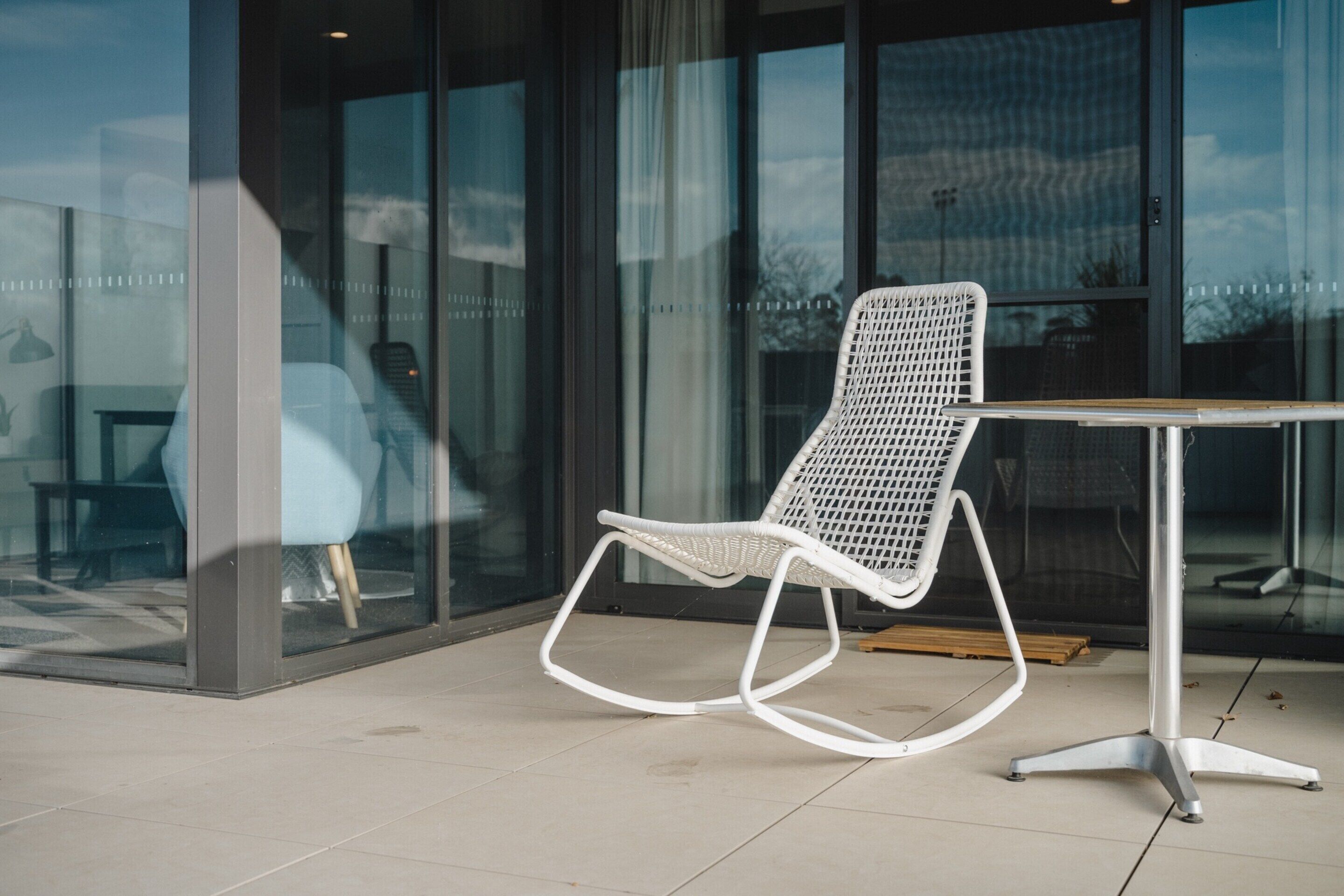 A white rocking chair with a metal frame and a table with a metal base, positioned on a tiled floor in front of a building with large glass windows.