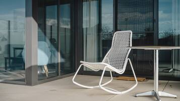 A white rocking chair with a metal frame and a table with a metal base, positioned on a tiled floor in front of a building with large glass windows.