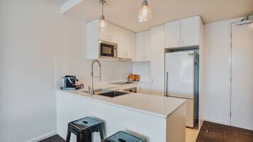 A modern kitchen with white cabinetry, a stainless steel refrigerator, a microwave, and two bar stools.
