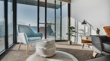 A modern living room with a large sliding glass door leading to a balcony, a blue armchair, a round ottoman, a black leather sofa, and a floor lamp.