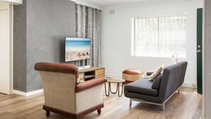 A living room with a television on a wooden stand, a leather armchair, a grey sofa, and a small round table. - Suburban Two-bed Apartment With Parking and Patio (Sydney)