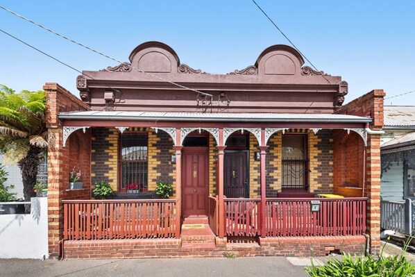 A two-story brick building with a red front door, a balcony with a metal railing, and a decorative cornice.
