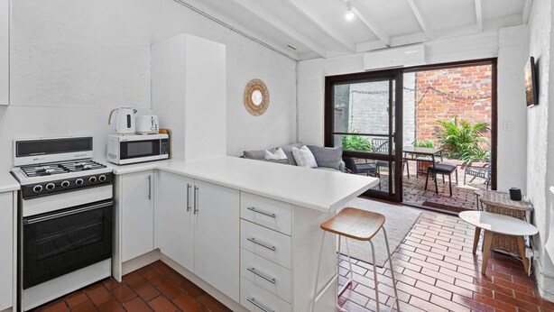 A compact kitchen with white cabinets, a stove, and a microwave. There's a small seating area with a table and chairs outside, a balcony with a view of a brick building, and a round mirror on the wall.