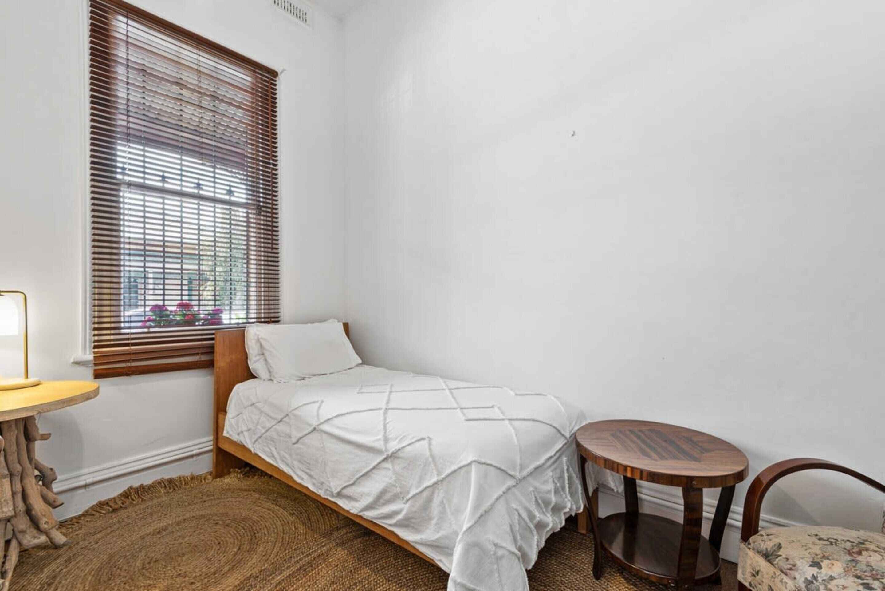 Un dormitorio pequeño y ordenado con una cama individual, una mesa redonda de madera, una silla y un ventanal con persianas.