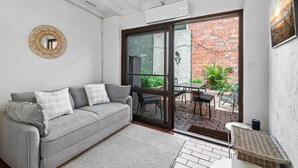 A modern living room with a grey sofa, a large sliding glass door leading to an outdoor patio with a brick wall and greenery.
