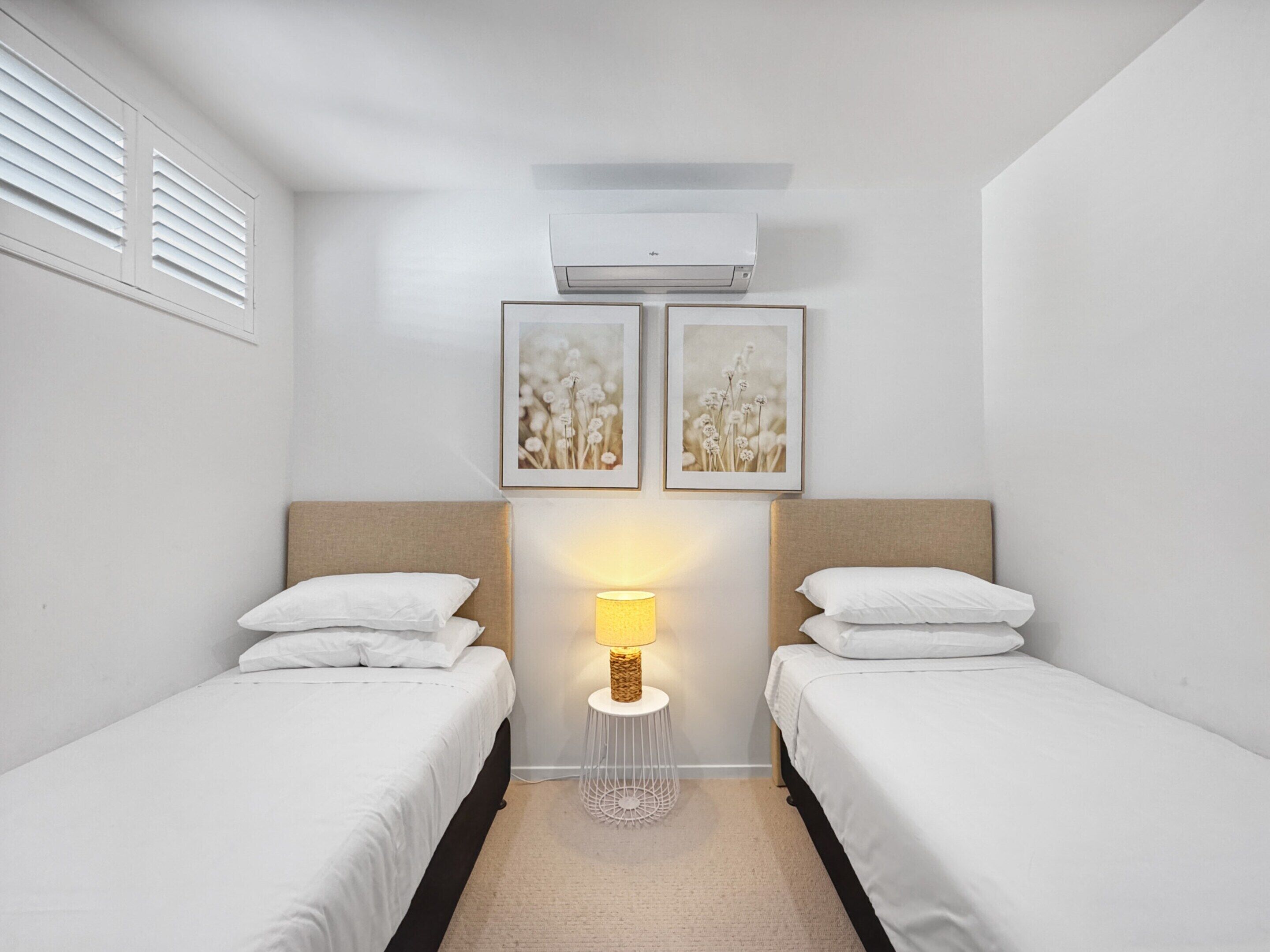 A bedroom with two beds, a wall-mounted air conditioner, and two framed pictures above the beds.