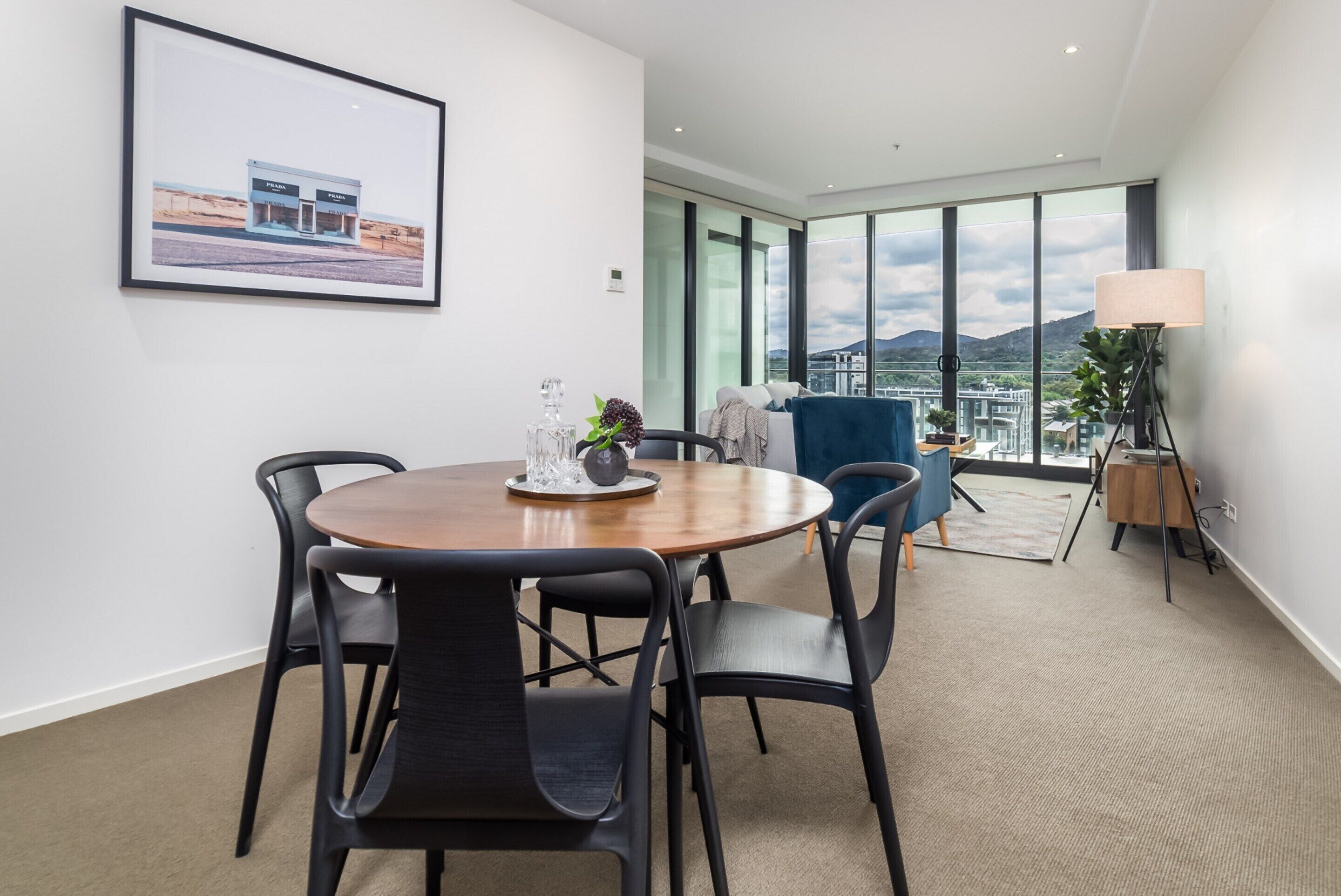 A modern hotel room with a round dining table, black chairs, a large window with a city view, and a framed picture on the wall.