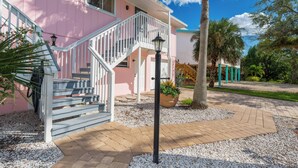 A pink house with a white staircase leading to a porch, surrounded by gravel and plants.