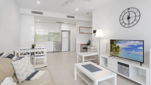 A modern living room with a sofa, coffee table, TV, and a kitchen in the background. - Beautiful Unit With Gym and Pool Near Museums (Brisbane)