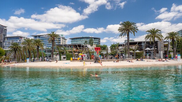 A beach with clear turquoise water, palm trees, and modern buildings in the background. - Beautiful Unit With Gym and Pool Near Museums (Brisbane)