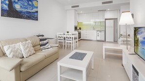 A modern living room with a beige sofa, a white coffee table, and a dining area with white chairs. A kitchen with white cabinets and a refrigerator is visible in the background. - Beautiful Unit With Gym and Pool Near Museums (Brisbane)