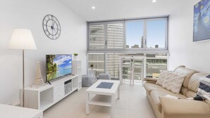 A modern living room with a large window, a flat-screen TV, a sofa, and a coffee table. - Beautiful Unit With Gym and Pool Near Museums (Brisbane)
