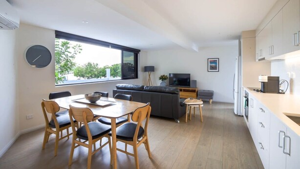Eine moderne Küche mit weißen Schränken, ein Essbereich mit einem Holztisch und -stühlen, ein graues Sofa und ein großes Fenster für natürliches Licht.