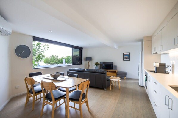 A modern kitchen with white cabinets, a dining area with a wooden table and chairs, a grey sofa, and a TV in the background. - 2-bedroom Apartment in the Heart of Batemans Bay (Batemans Bay)