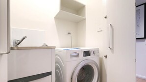 Kompakti pyykinhuoltotila, jossa on Bosch-merkkinen pesukone ja -kuivausrumpu, pieni hylly kuivausrummun yläpuolella sekä ovi, joka johtaa toiseen huoneeseen.