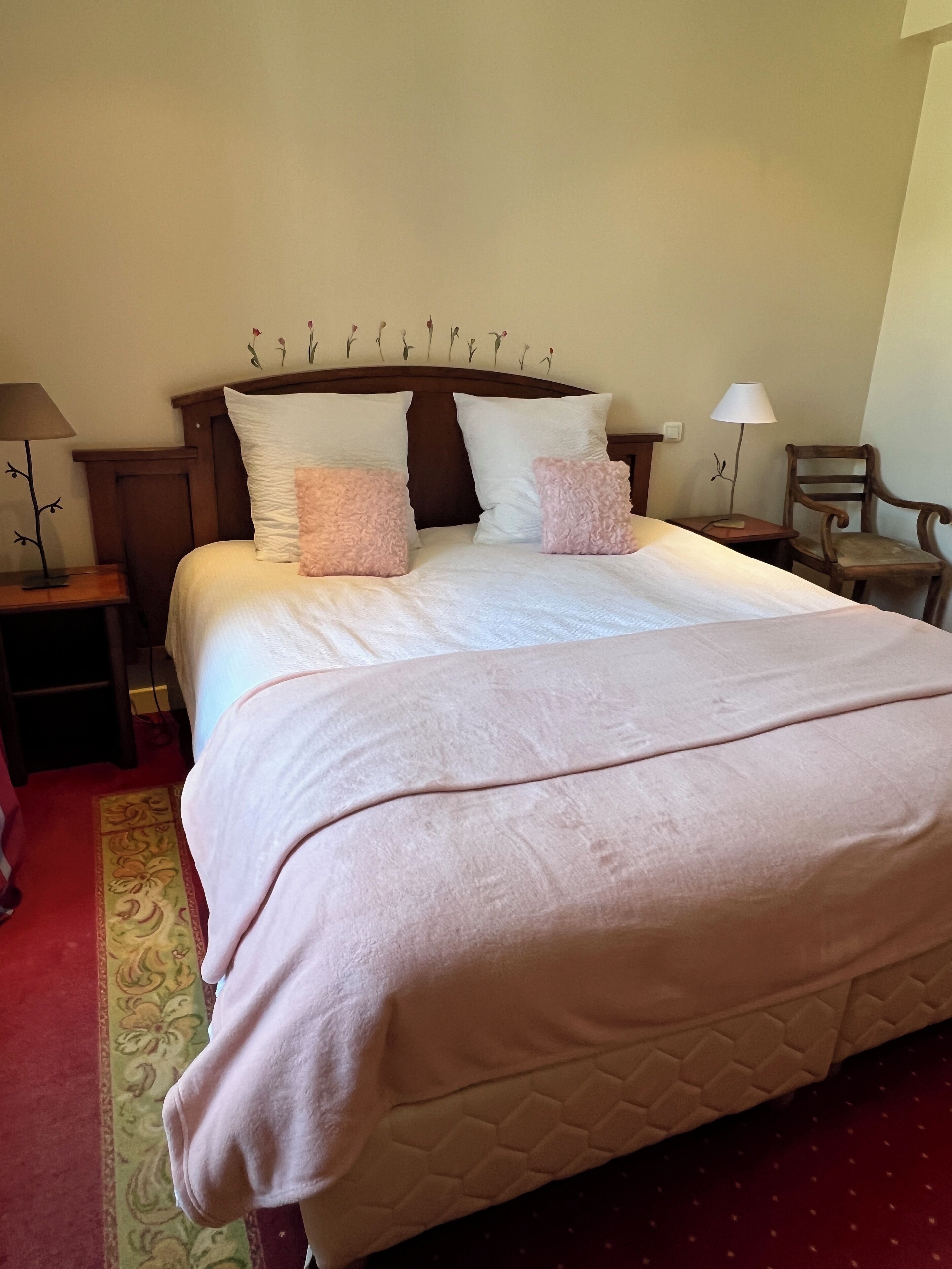 A neatly made bed with a quilted cover, two bedside tables with lamps, a chair, and a decorative wall piece above the bed.