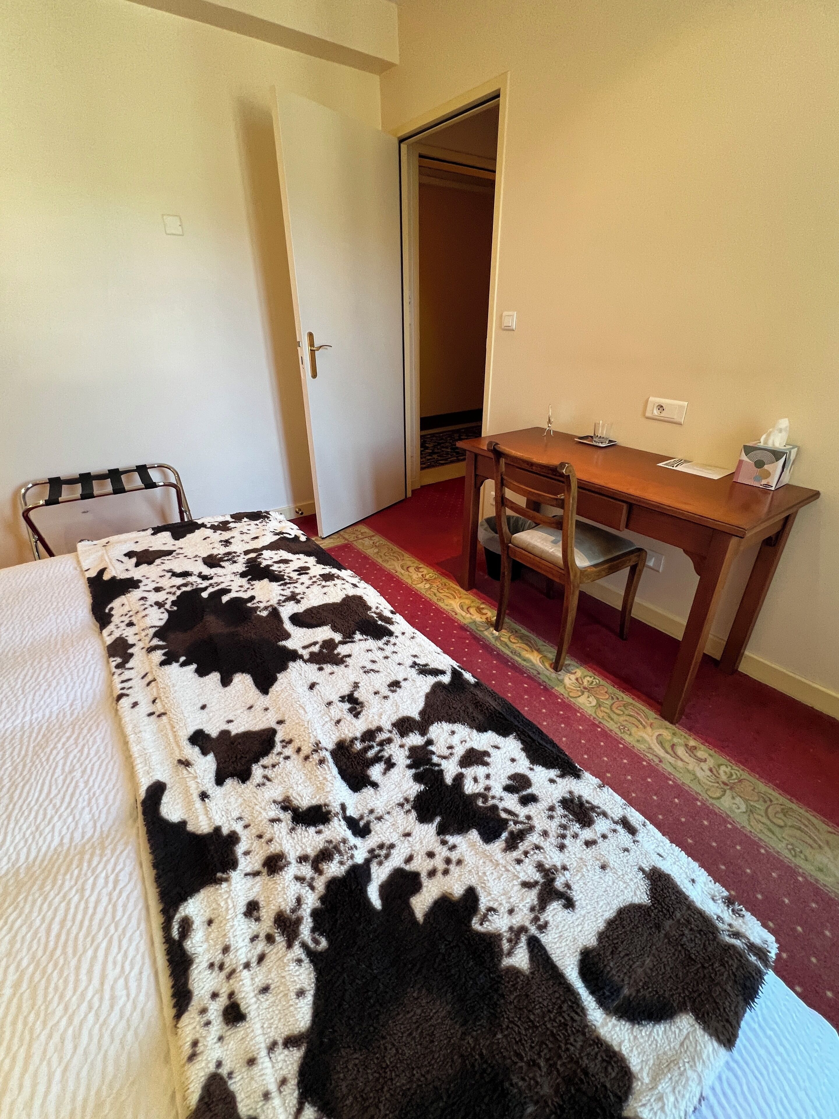 A bedroom with a bed, a desk, and a chair. There is a door leading to another room.