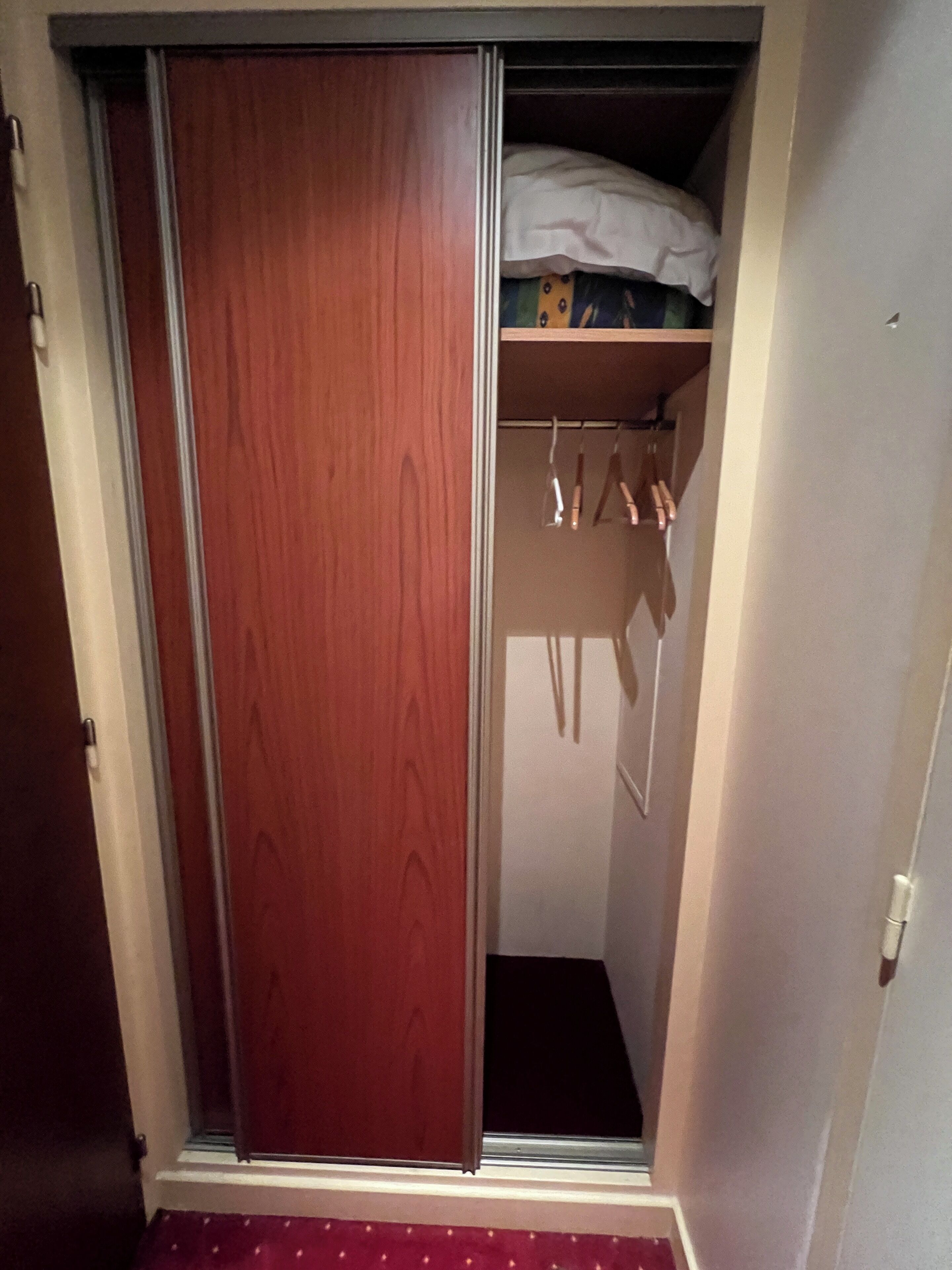 Une armoire en bois dotée d’une porte coulissante, d’une étagère au-dessus où sont pliés des vêtements, et d’une tringle pour suspendre les vêtements.