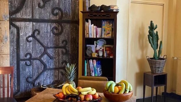 A dining area with a wooden table, chairs, a fruit bowl, a bookshelf, and a cactus plant. - Home Le Soft (Vic-sur-Cère)