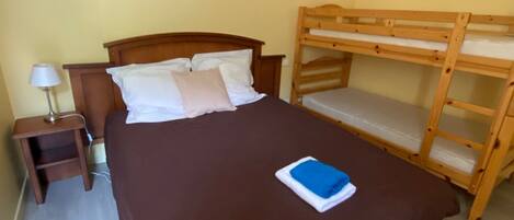Una cama individual con una colcha marrón, una mesita de noche con lámpara, y una cama litera a la derecha.