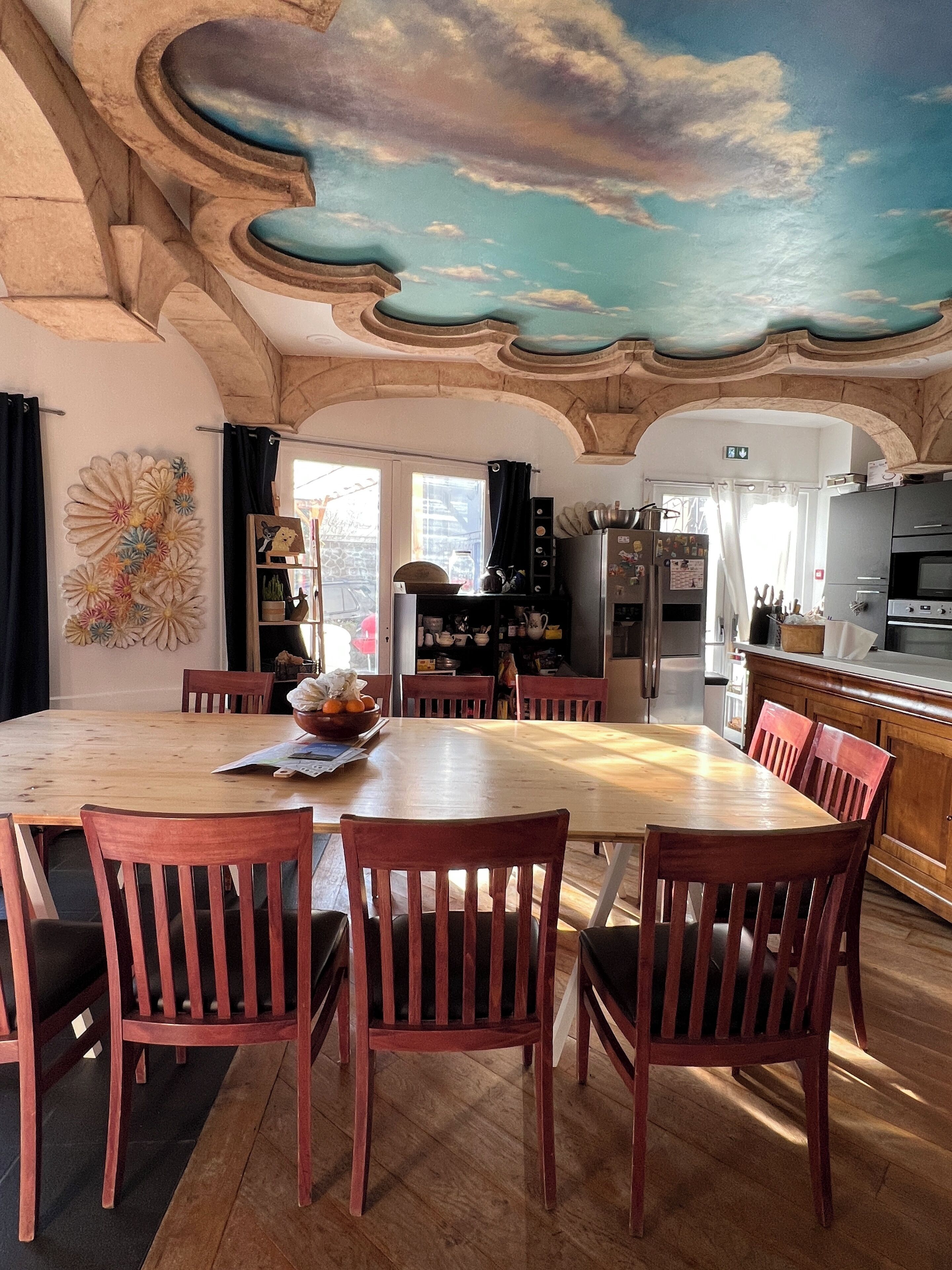 A dining area with a wooden table and chairs, a decorative ceiling mural, and a kitchen in the background.