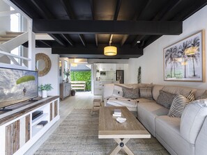 A modern living room with a sofa, coffee table, and television. There is a kitchen visible through an open door. - Beachside 2-bed House With Pool, Courtyard & BBQ (Sunshine Coast)