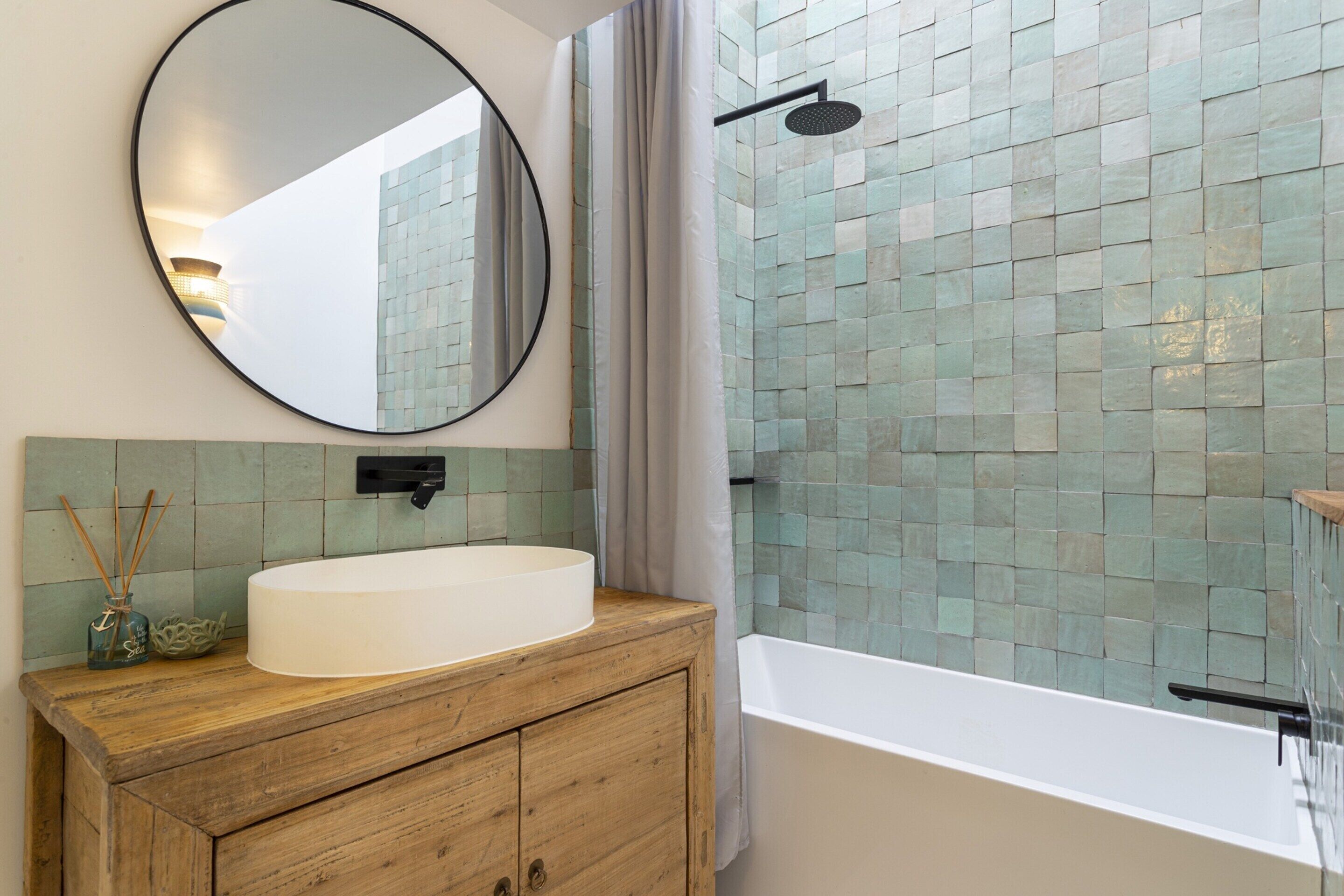 A bathroom with a round mirror, a wooden vanity, a white sink, a shower with a tiled wall, and a bathtub.