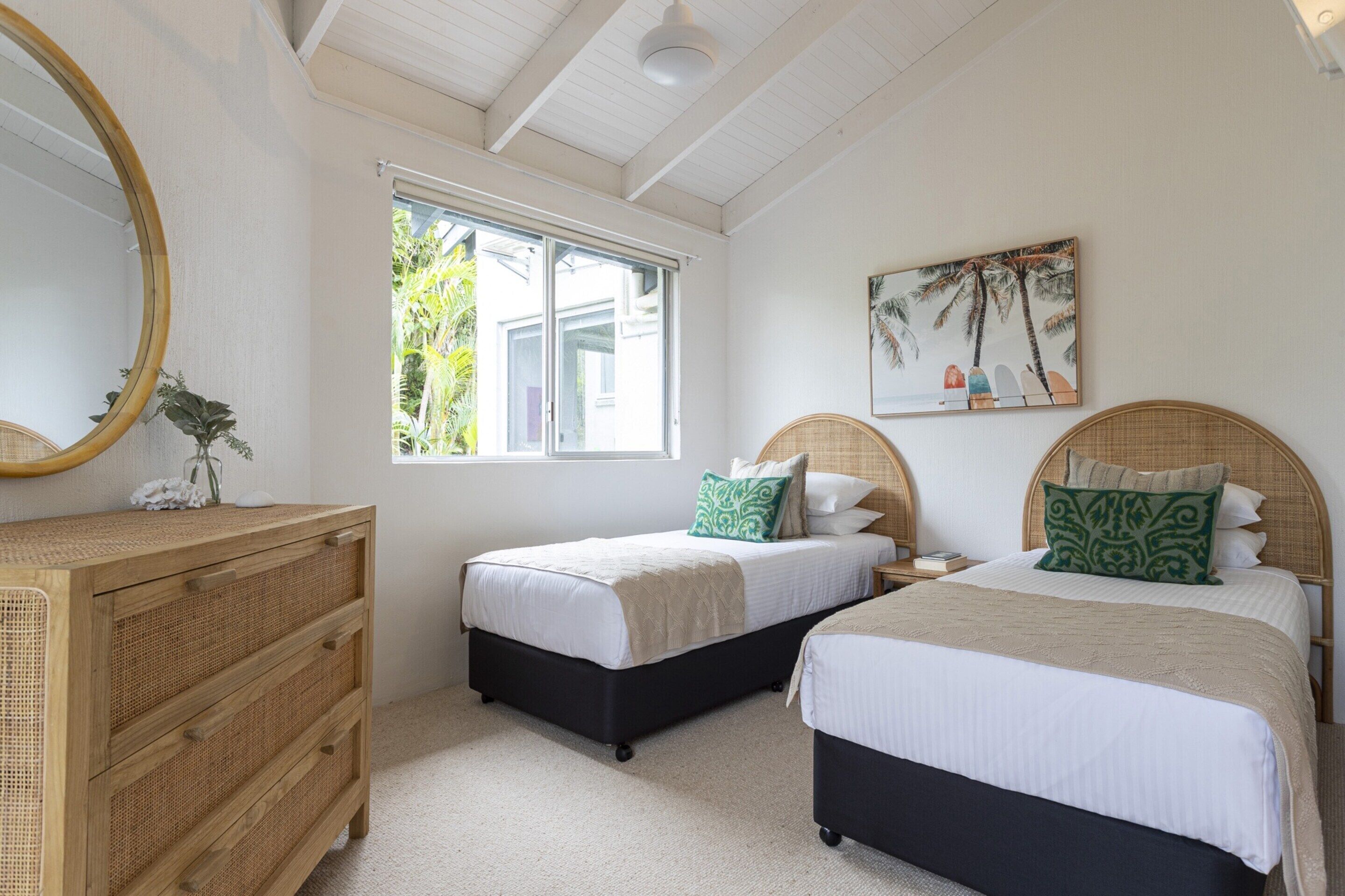 A bedroom with two beds, a dresser, a mirror, and a window with a view of greenery.
